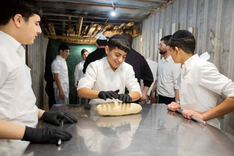 Teenage boys in white shirts and black gloves shape matzah dough on a long steel table; one of them is smiling.