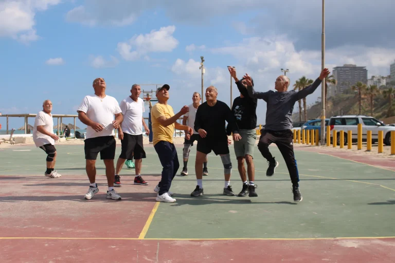 A group of older men on an outdoor sports court by the sea look up and reach for a ball that is outside the picture; one of them jumps.