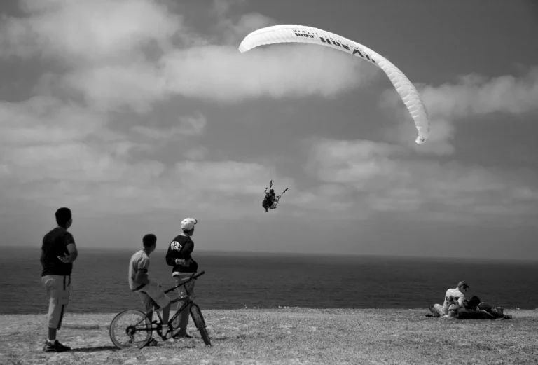 Black-and-white photograph: a paraglider flies above the sea while three people, one with a bicycle, watch from the clifftop.