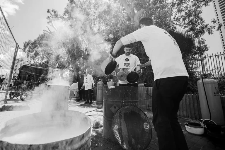 Black-and-white photograph: a man in a white shirt lowers cookware into a steaming vat outdoors while others wait behind him.