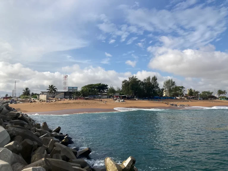 A calm bay with a sandy beach, trees and low buildings, and dark rocks in the foreground.