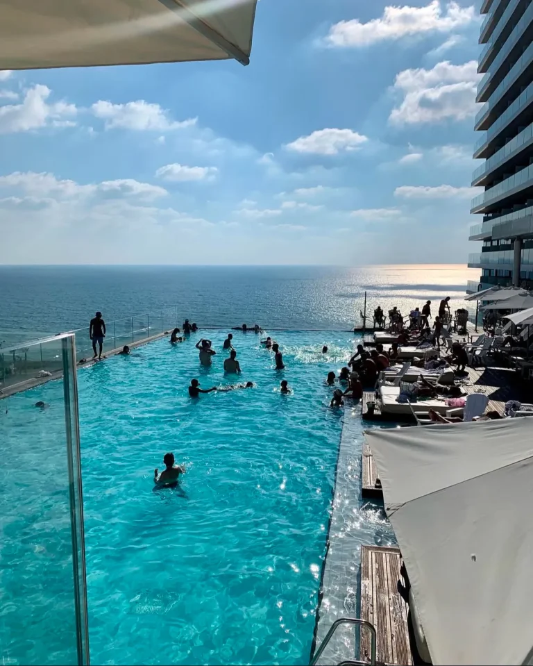 View from above of a turquoise hotel pool with swimmers, with the sea and a cloudy sky beyond it.