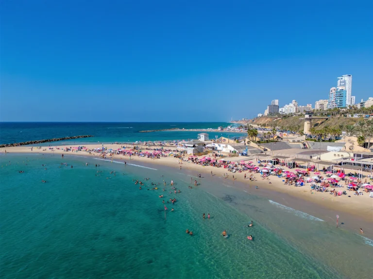 Turquoise sea and a sandy beach crowded with pink umbrellas, with the towers of Netanya on the cliff behind.